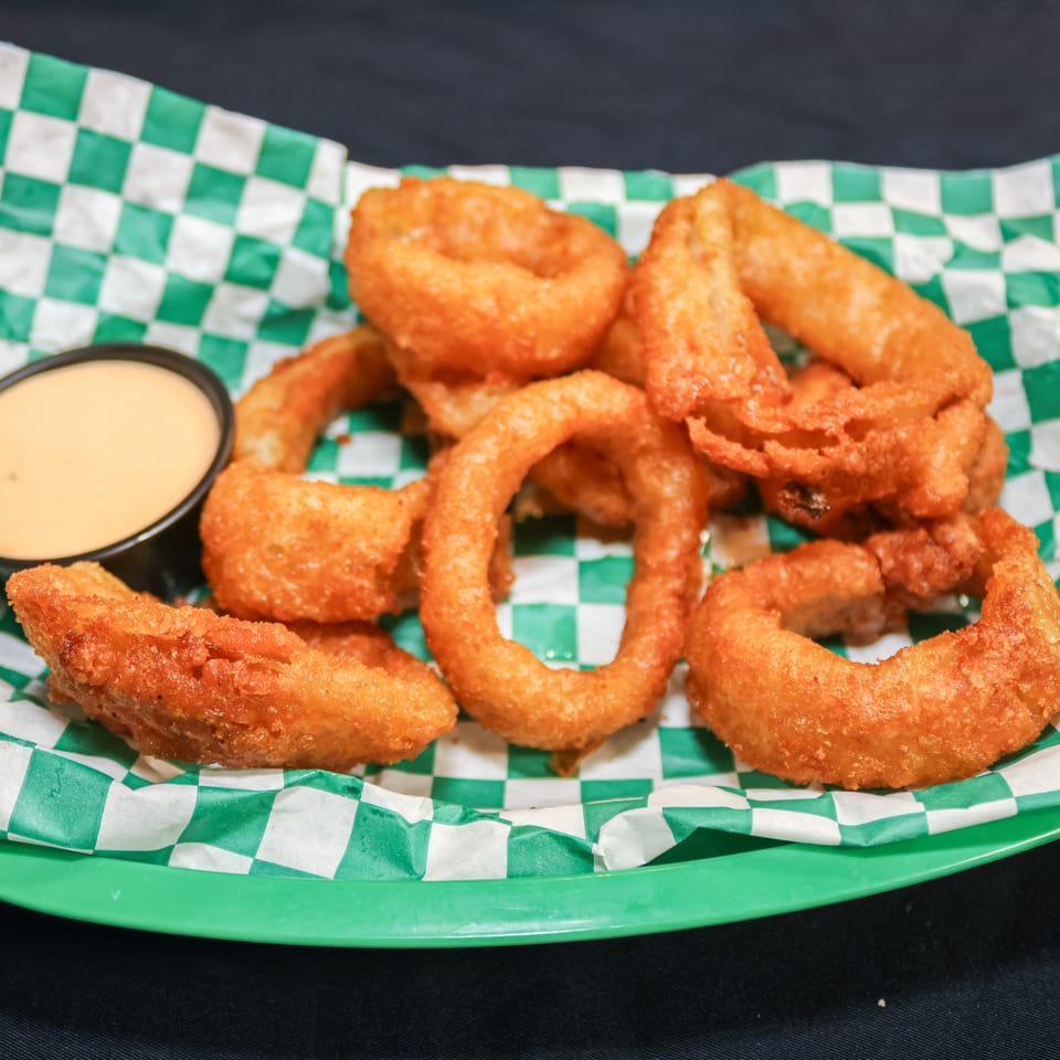 A basket of golden-brown onion rings with a side of dipping sauce on a green and white checkered paper.