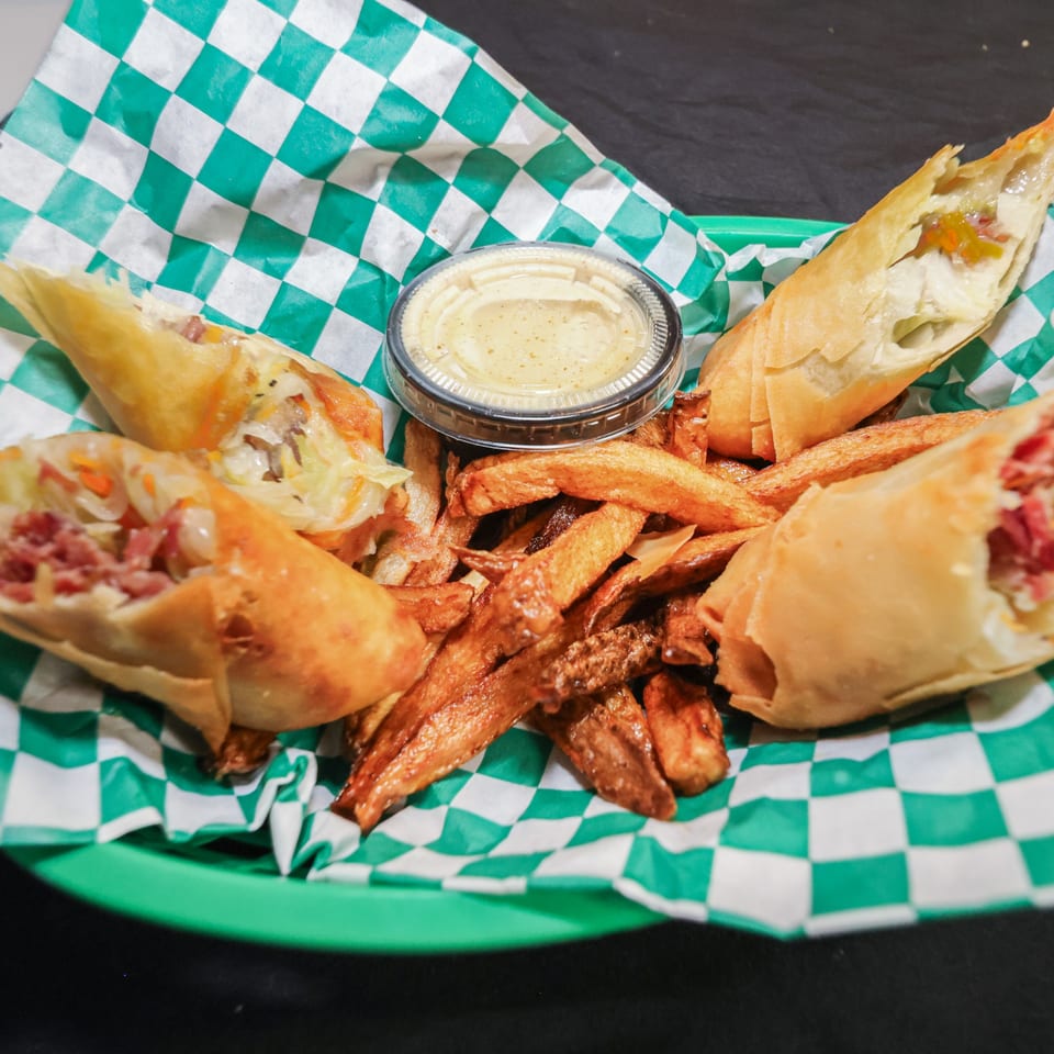 Four crispy egg rolls, two cut open to show fillings, with fries and dipping sauce in a green basket.