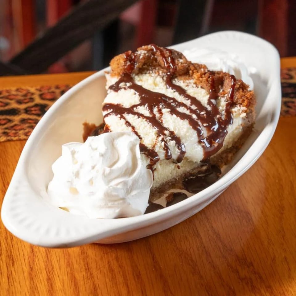 A slice of cheesecake with chocolate drizzle and whipped cream in a white dish on a wooden table.